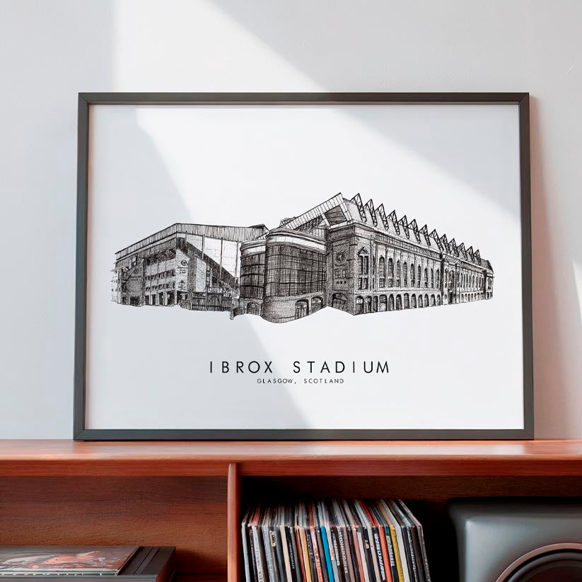 A framed black and white architectural art print of Ibrox Stadium in Edinburgh, Scotland, displayed on a wooden shelf against a light gray wall.