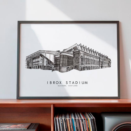 A framed black and white architectural art print of Ibrox Stadium in Edinburgh, Scotland, displayed on a wooden shelf against a light gray wall.